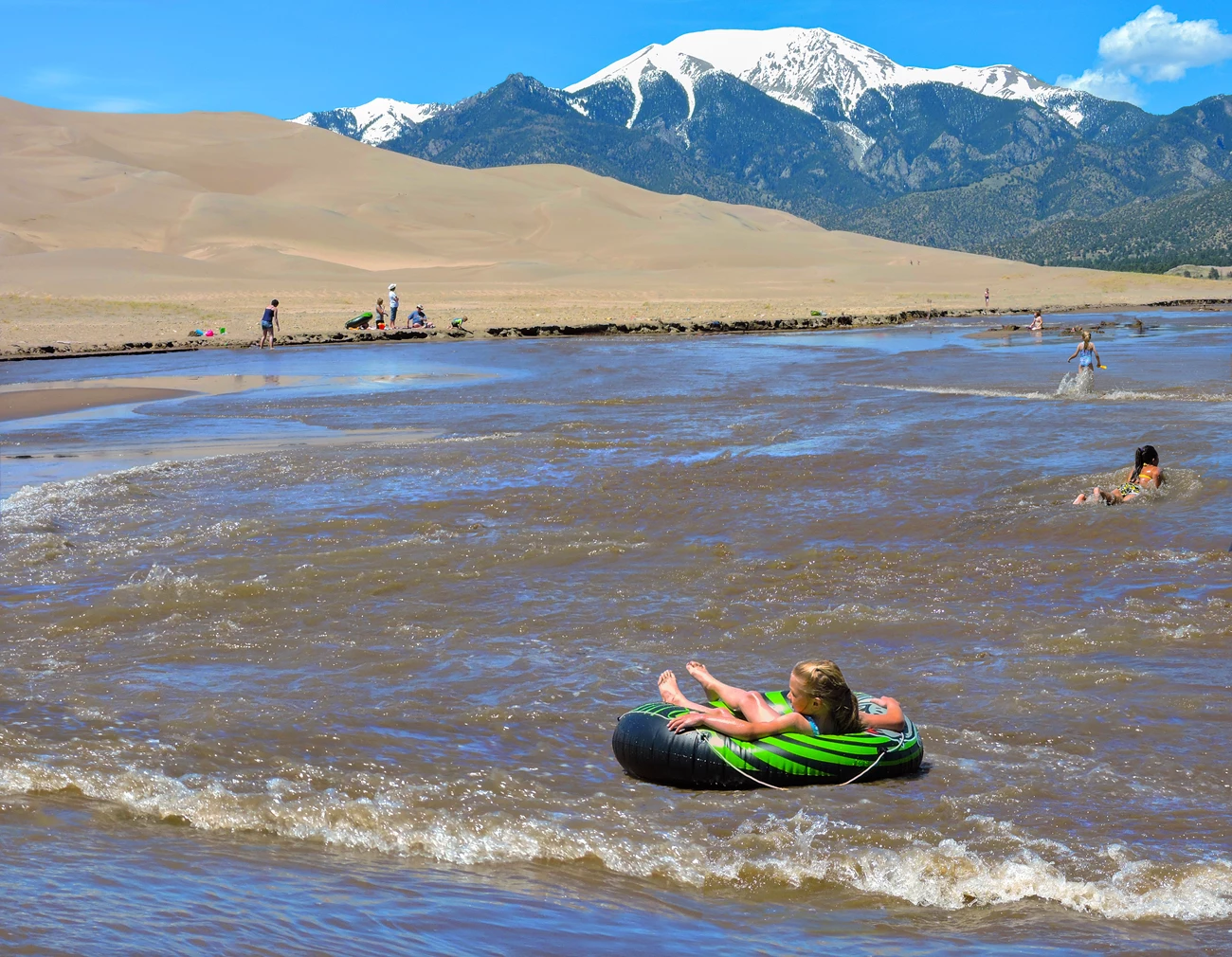 Visitors wading through shallow Medano Creek at the edge of the Great Sand Dunes with tan dunes rising behind them under a bright Colorado sky, documentary travel photo