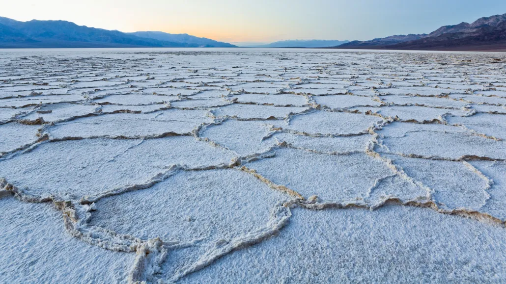 Wide view of Badwater Basin salt flats showing bright white polygon salt patterns with dark mountains in the distance under clear sky, realistic travel photography