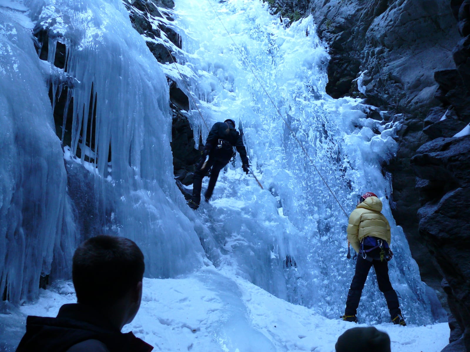 Zapata Falls pouring between narrow rock walls with a visitor standing in shallow stream water looking toward the waterfall in shaded canyon light
